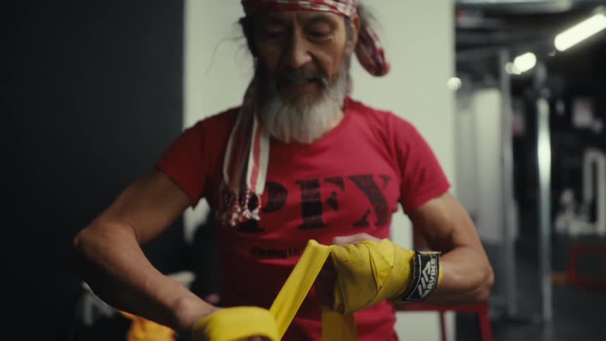 Elderly man bandaging his hands with a yellow bandage inside a gym