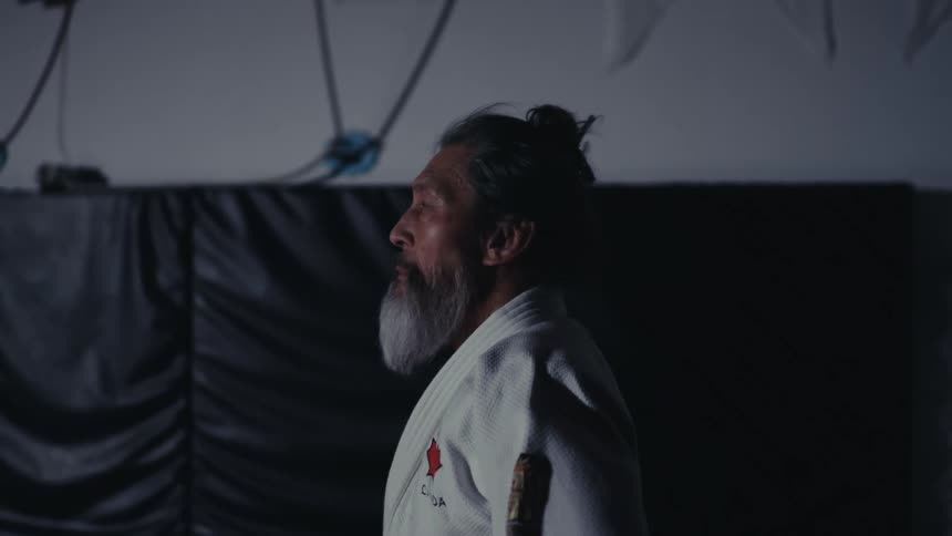 A man in a martial arts uniform stands in the dark gym