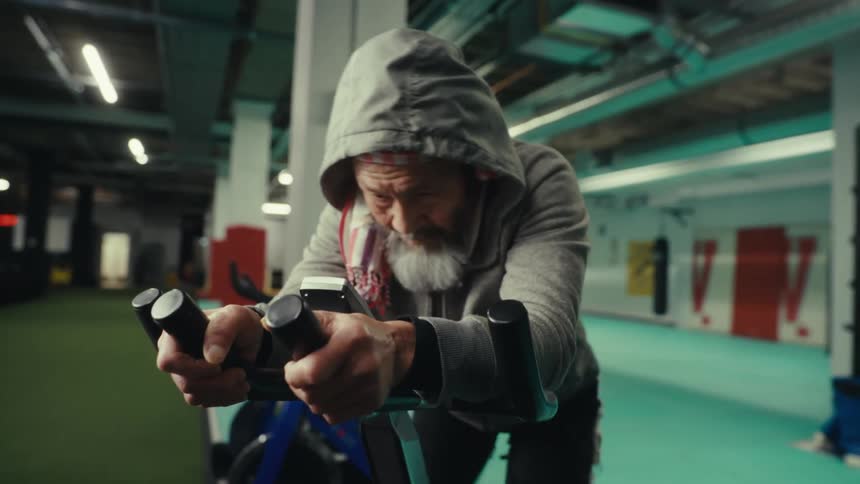 An old man in a hoodie rides an exercise machine in a gym