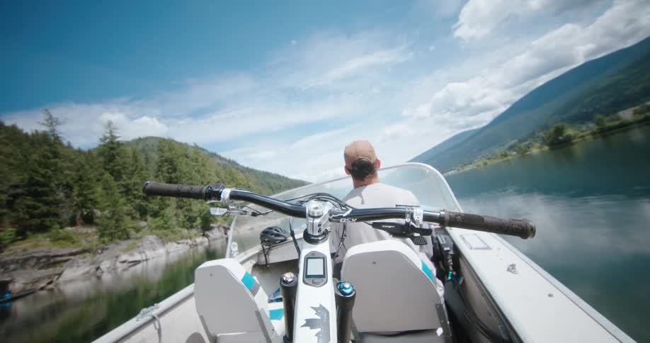 Man drives a boat across the lake between two mountains as seen from the perspective of his mountain bike