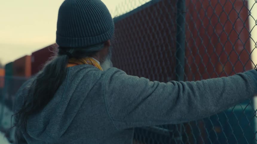 An old man with a hat and gloves stands with his hand resting on a metal railing watching a train passing by
