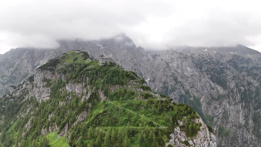 Switchback leads through the forest on a mountainside from a house on a ledge to a building on a ridge overlooking the valley on a misty and cloudy day
