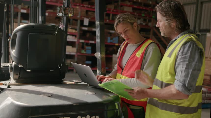 Two male coworkers consult a laptop and a clipboard while working