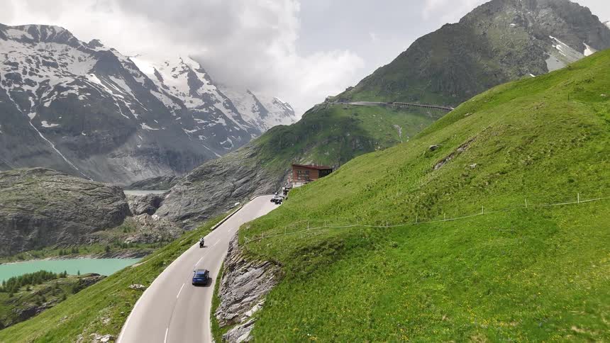 A blue car driving on a winding mountain road surrounded by lush green slopes and snow capped peaks in the background