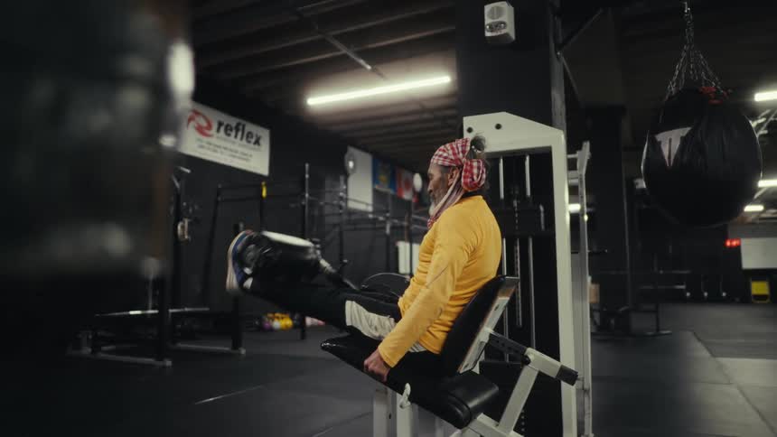 A man with a cloth on his head and married in yellow is doing weight lifting with his legs in a gym