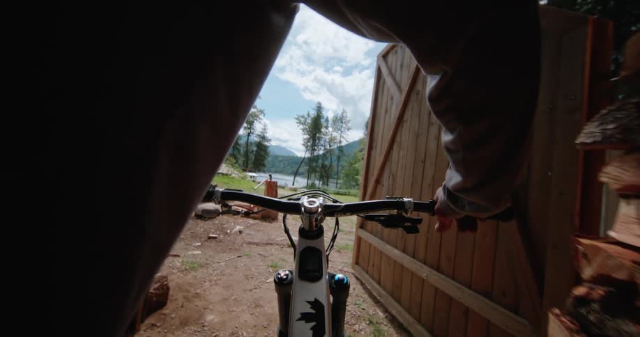Male cyclist rides bicycle out of wood shed and rides towards the lake at the bottom of the hill
