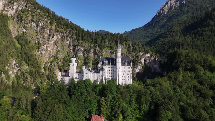 Aerial view of a white castle on a hill surrounded by vegetation and in the background of imposing mountains