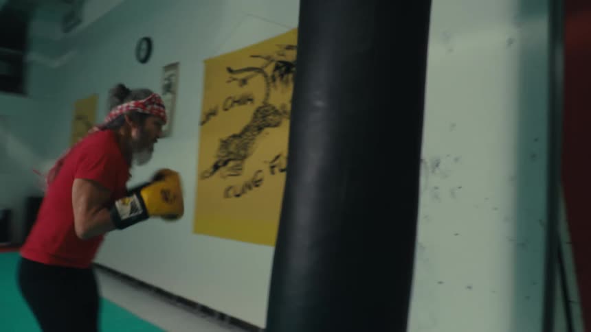 A bearded man wearing boxing gloves practices punching on a heavy bag in a gym