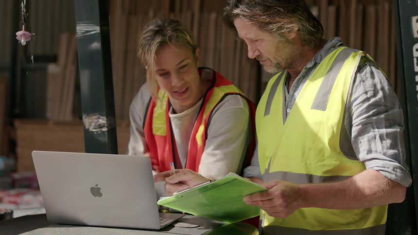 A man and a woman in builders clothes are talking while looking at a laptop on a table
