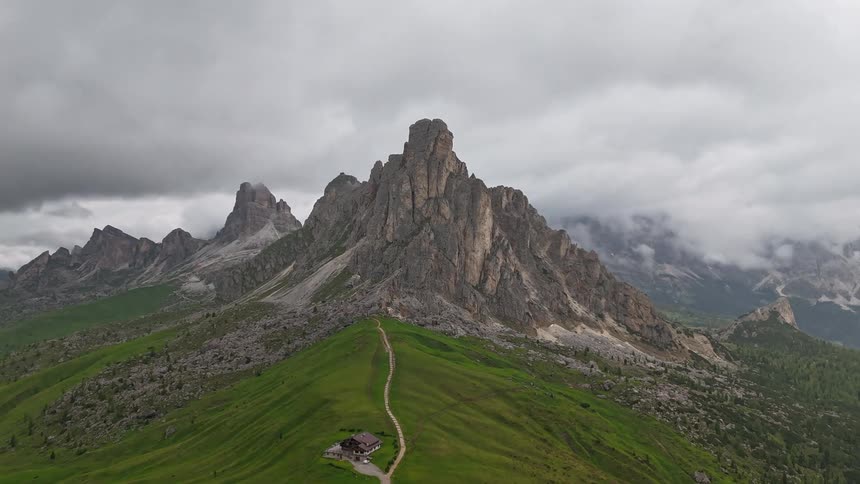 Cars visit a large building built near a tall mountain range as dark clouds move overhead