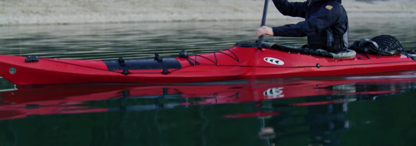 Man paddling a red kayak in calm waters with a shoreline in the background