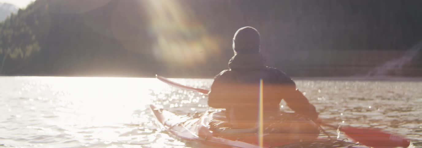 Sun glares down on man paddling red kayak off a forest covered mountainous coast