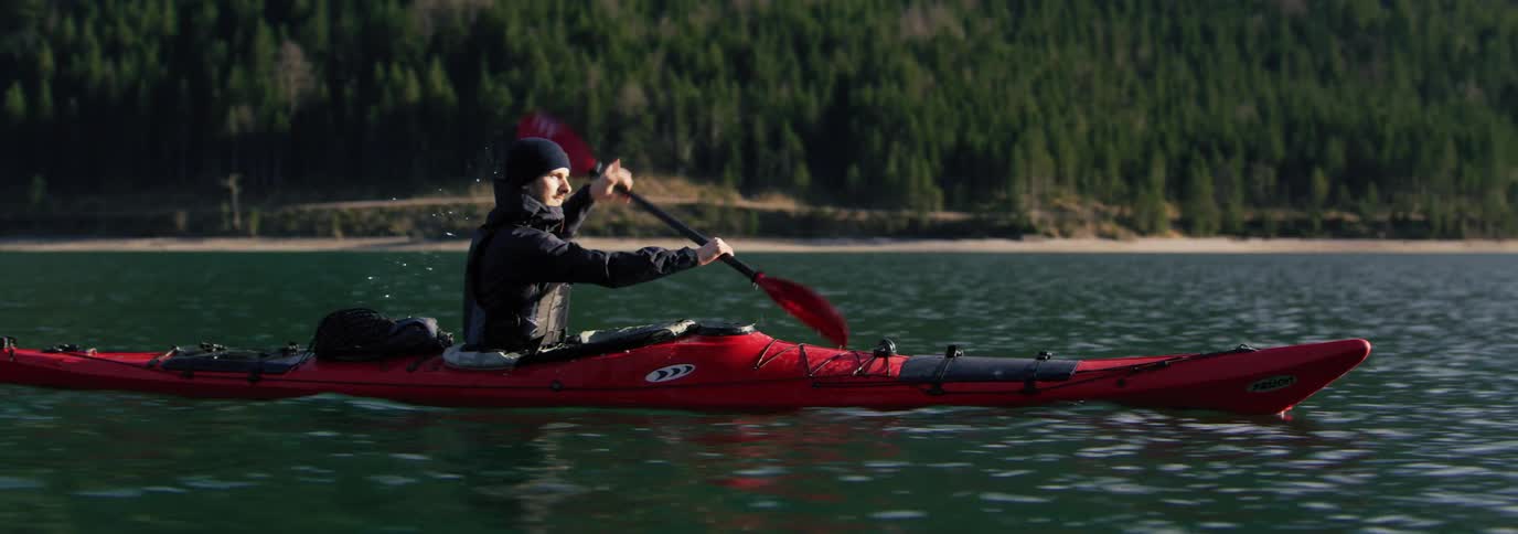 Male kayaker paddles a red kayak through the water with a forested mountainous shore in the background