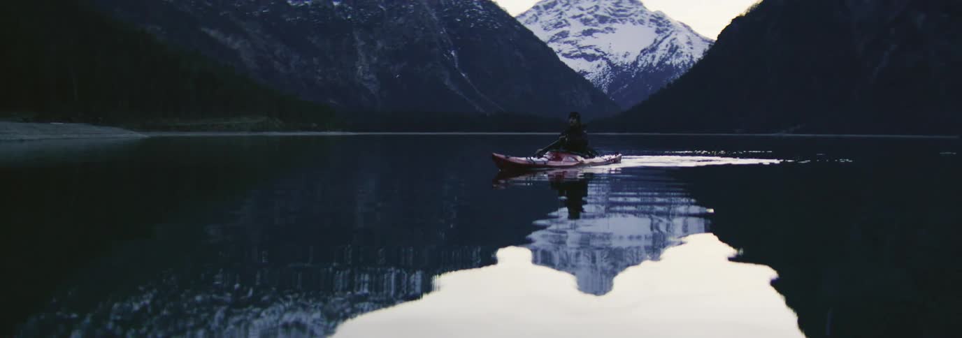 Person paddling a red kayak in calm waters with snowy mountains in the background