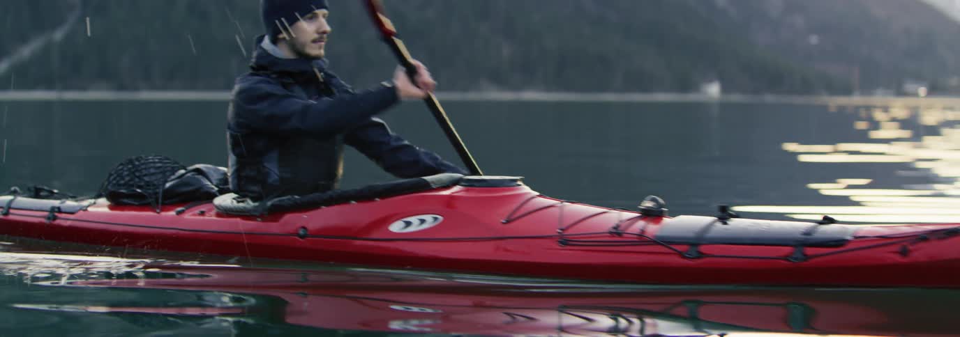 Man paddling a red kayak on a large body of water with mountains and trees in the background