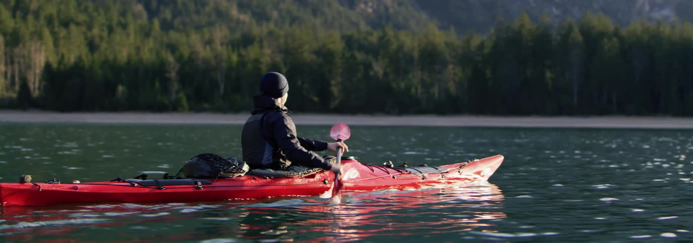 Man paddling a red kayak in calm waters with a coastline of mountains and trees in the background