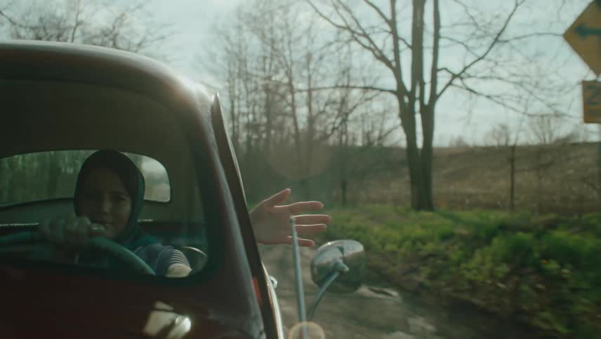A woman smiles as she drives down a rural road with her hand out of the car window