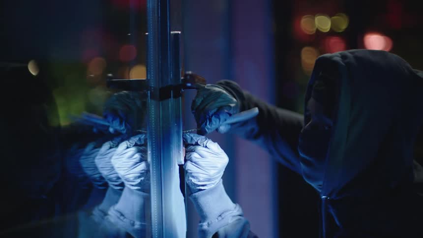 A person with their face partially covered using a tool to pry open a glass door at night