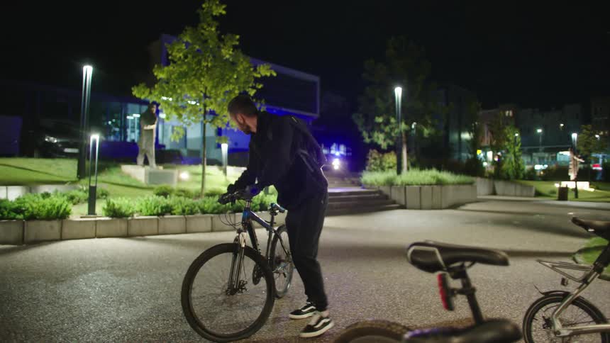 Two men in black clothes stealing bicycles from a bike shop at night and one of them drops the bike on the ground and runs