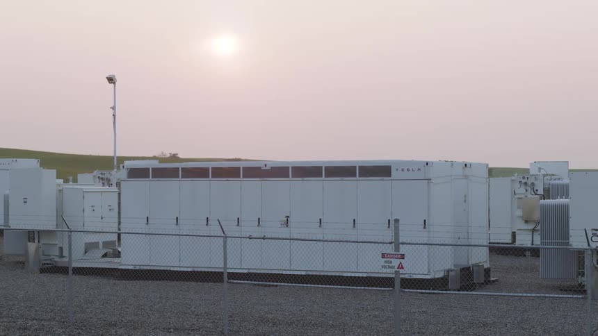 A large white industrial megapack battery unit inside a fenced facility during sunset