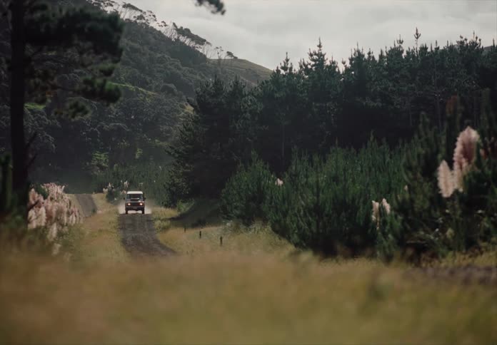 Vehicle creates dust while moving along a rural highway passing forested hills on an overcast day