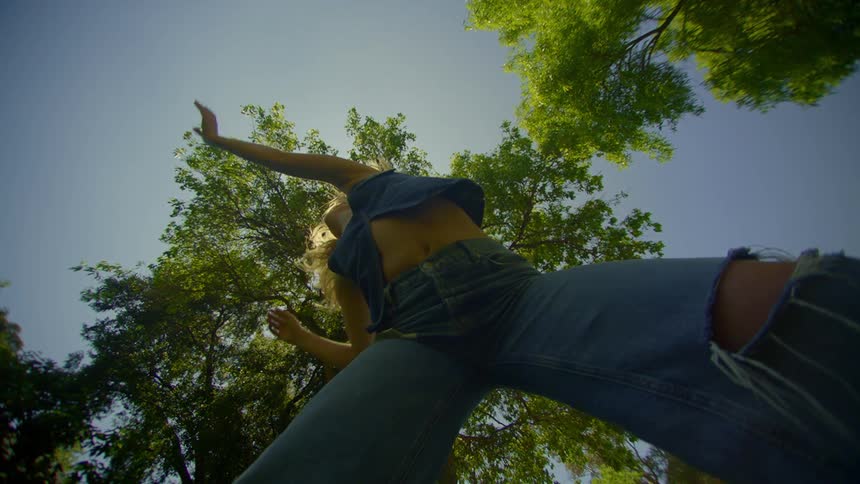 A woman dances outdoors wearing denim with sunlight filtering through tree leaves