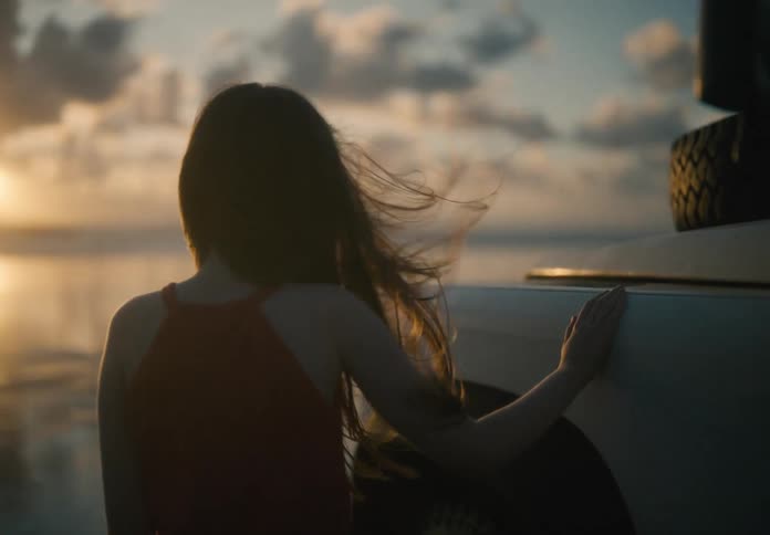 A girl with long hair standing placing her hand on a truck next to the water at sunset