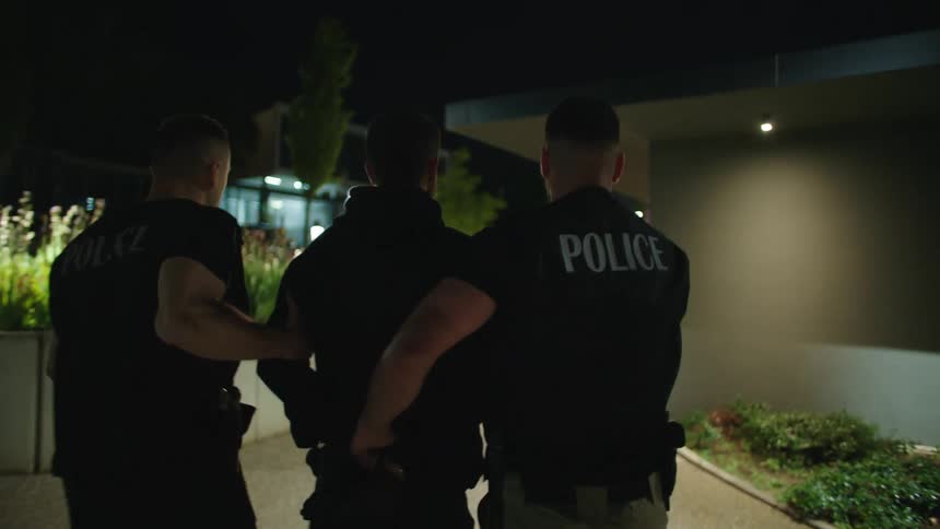 Two policeman handcuff a suspect and walk him toward their police car passing a building at night