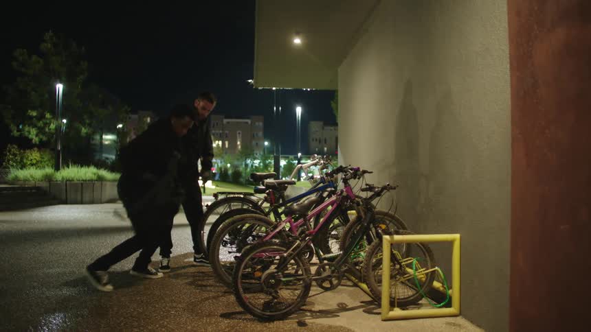 Two men in black stealing a bicycle from an outdoor establishment with pliers