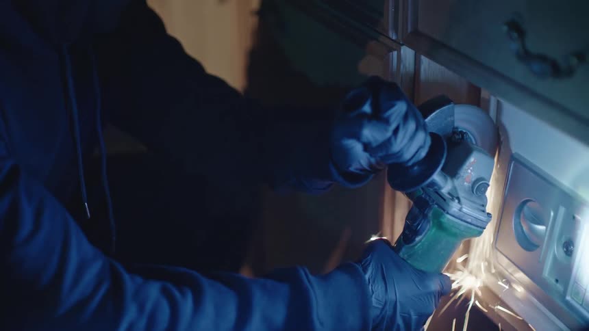 Two thieves one with a flashlight and the other with a saw destroying a safe in a dark environment