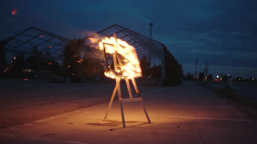 A wooden easel with a burning canvas stands in an empty outdoor space during twilight with sparks flying off the fire
