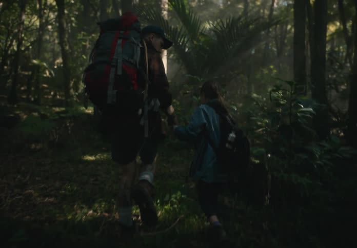 Two hikers one adult and one child trek through a dense forest with backpacks illuminated by filtered sunlight