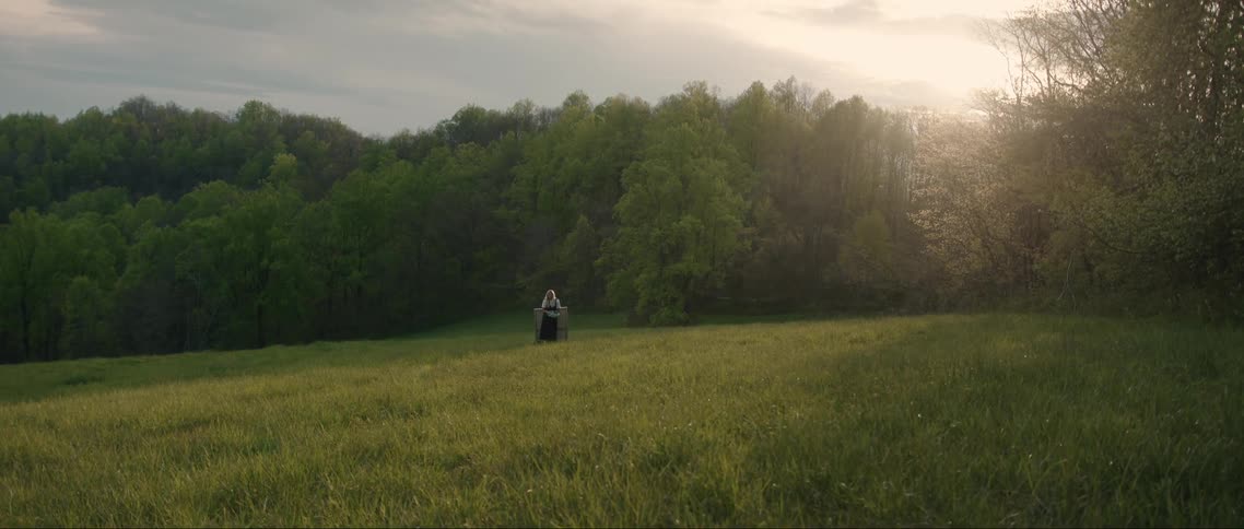 A woman with blond hair stand in the middle of a grassy area with trees in the background