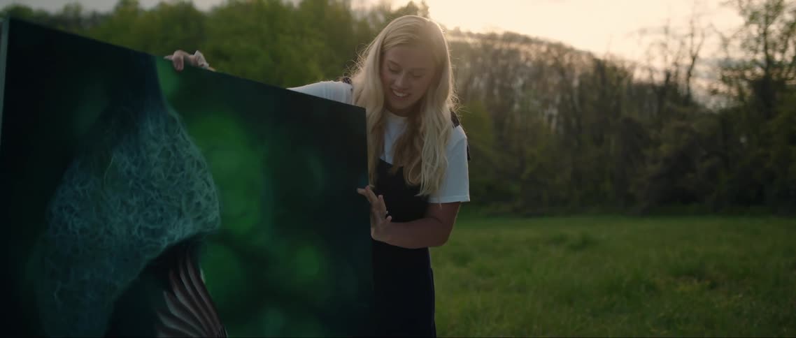 A young blonde woman handling a framed painting in a grassy field at sunset
