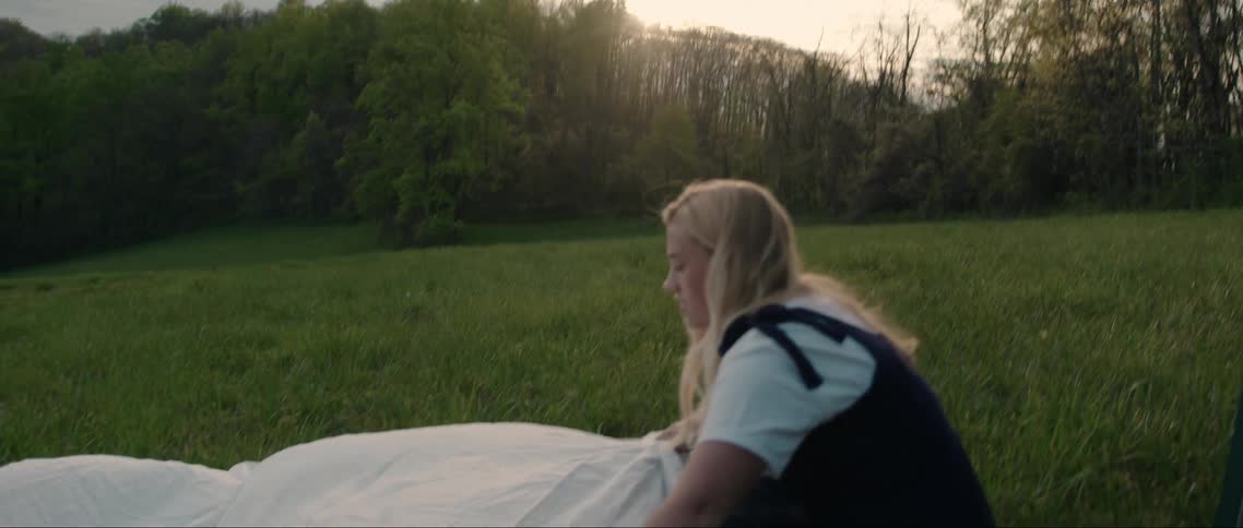 A young blonde woman is spreading a white cloth on a green grass