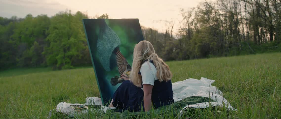 The blonde haired girl is sitting on a green and white blanket and has a canvas in front of her and she is painting a bird