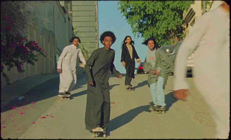 A group a kids are skateboarding down a road that has a small decline on itthe road is lined with cars and houses