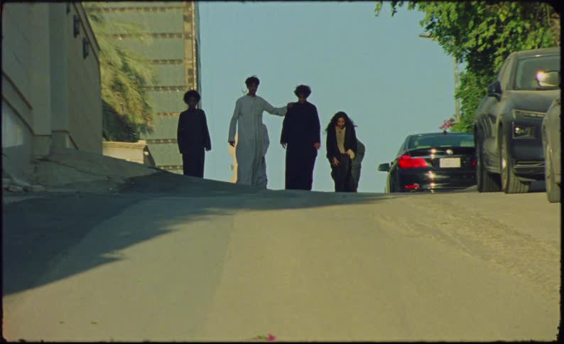 A group of people who are skateboarding on an asphalt street with some cars parked on the right