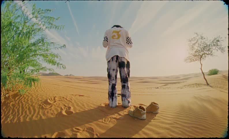 A man dressed in casual clothes kneeling in the desert praying stands under a blue sky
