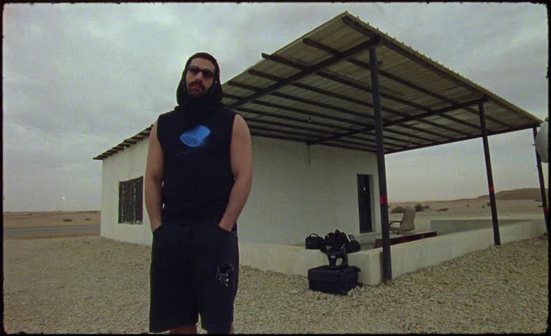 Man stands on arid terrain near a house with a large covered porch