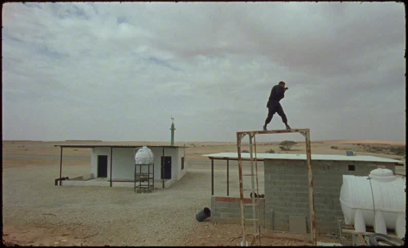 Male boxer balances on a narrow wooden frame while shadow boxing near buildings in the desert