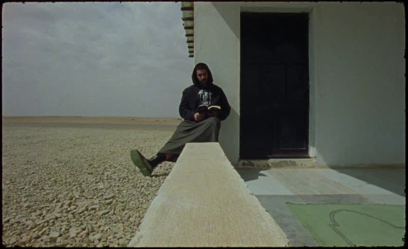 Man sits near the door of a building in arid terrain reading while a truck passes on the road in the background