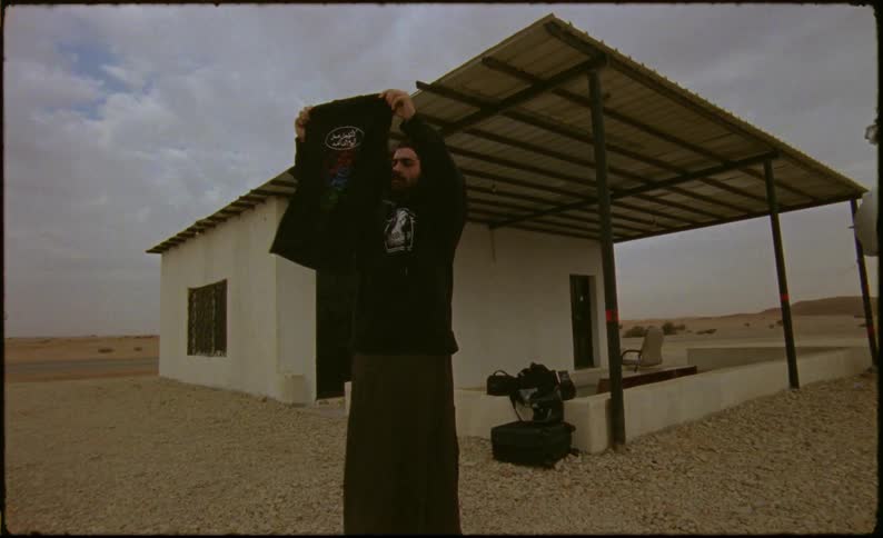Man in sweatshirt next to a small house looking at and lifting a black tshirt in a desert environment
