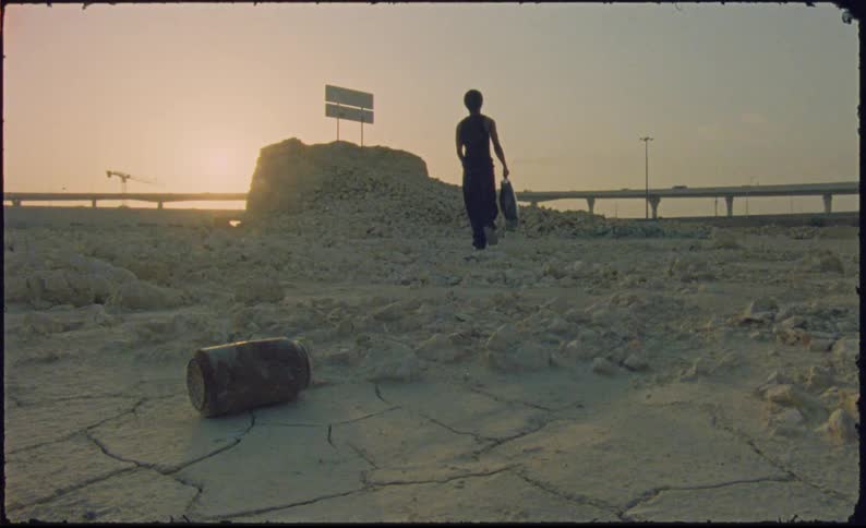 A person is walking with a bag in his hand on a sandy floor where there is a rusty bottle