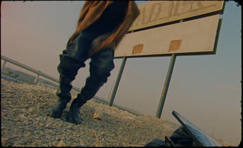 A model wearing boots and a brown jacket stands on a gravel mound near a large sign