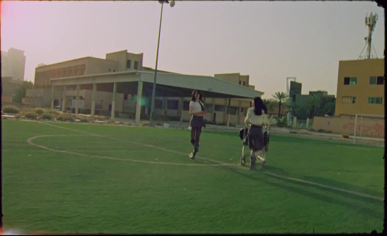 Two young university women in uniform running with a shopping cart with clothes inside an abandoned grass sports field