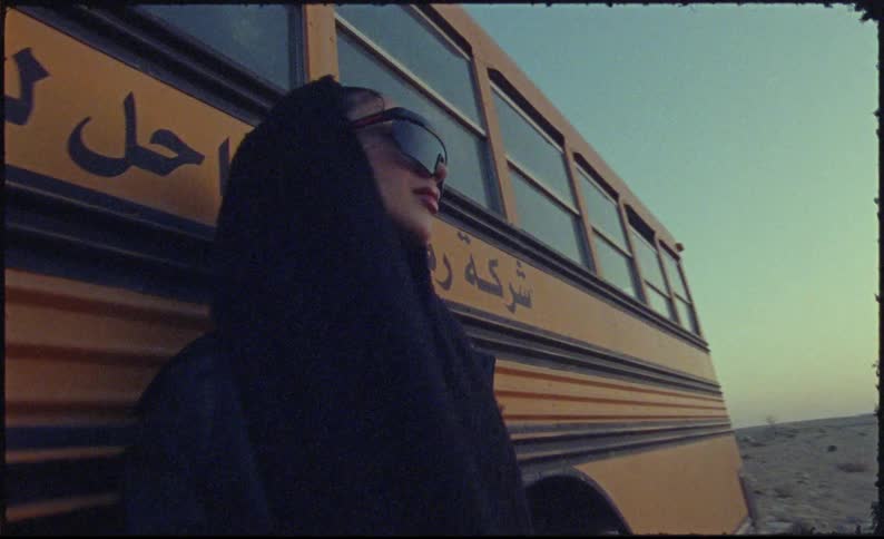 A woman with sunglasses and black clothes sits on a yellow bus in a desert region