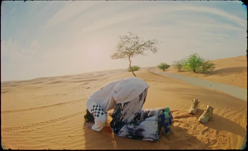 A man in a desert kneeling and bowing forward in a prayer position near a solitary tree with sparse foliage