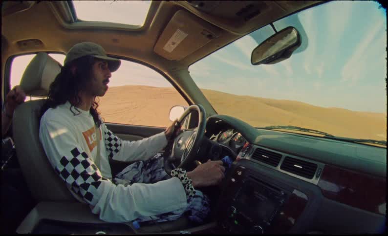 A man with a cap is in a car driving in a sandy area and skidding sideways