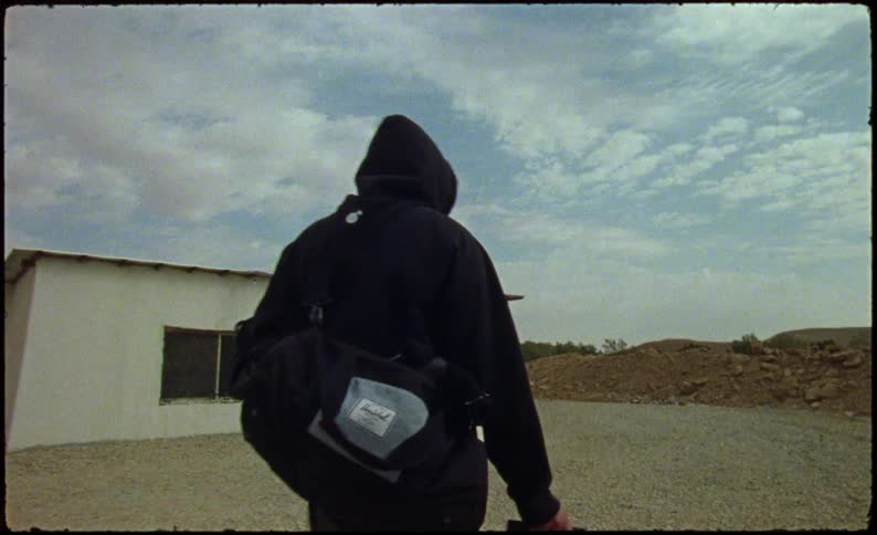 An individual in a black hoodie pulling a black suitcase with wheels near a solitary building in the desert